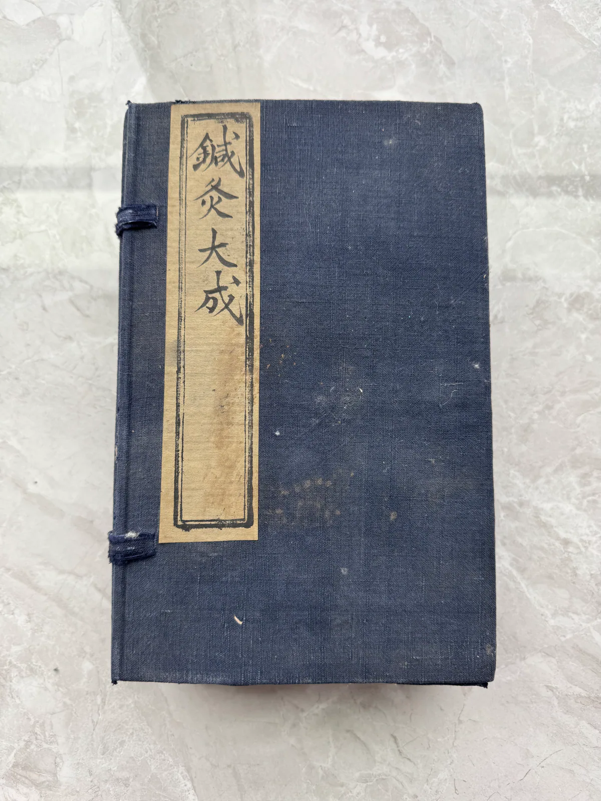 图片展示的是郑福兰堂珍藏版《针灸大成》书函。书函为深蓝色布质，边角有磨损痕迹，表面有明显折痕。书函正面有一张浅色纸条，纸条上用黑色毛笔书写着“针灸大成”四个字，纸条两侧有黑色边框装饰。书函被放置在浅色大理石纹理的桌面上，整体呈现出古朴典雅的外观。此图片与上下文紧密相关，直观呈现了文中提到的库房里被晒太阳的两件老物件之一。