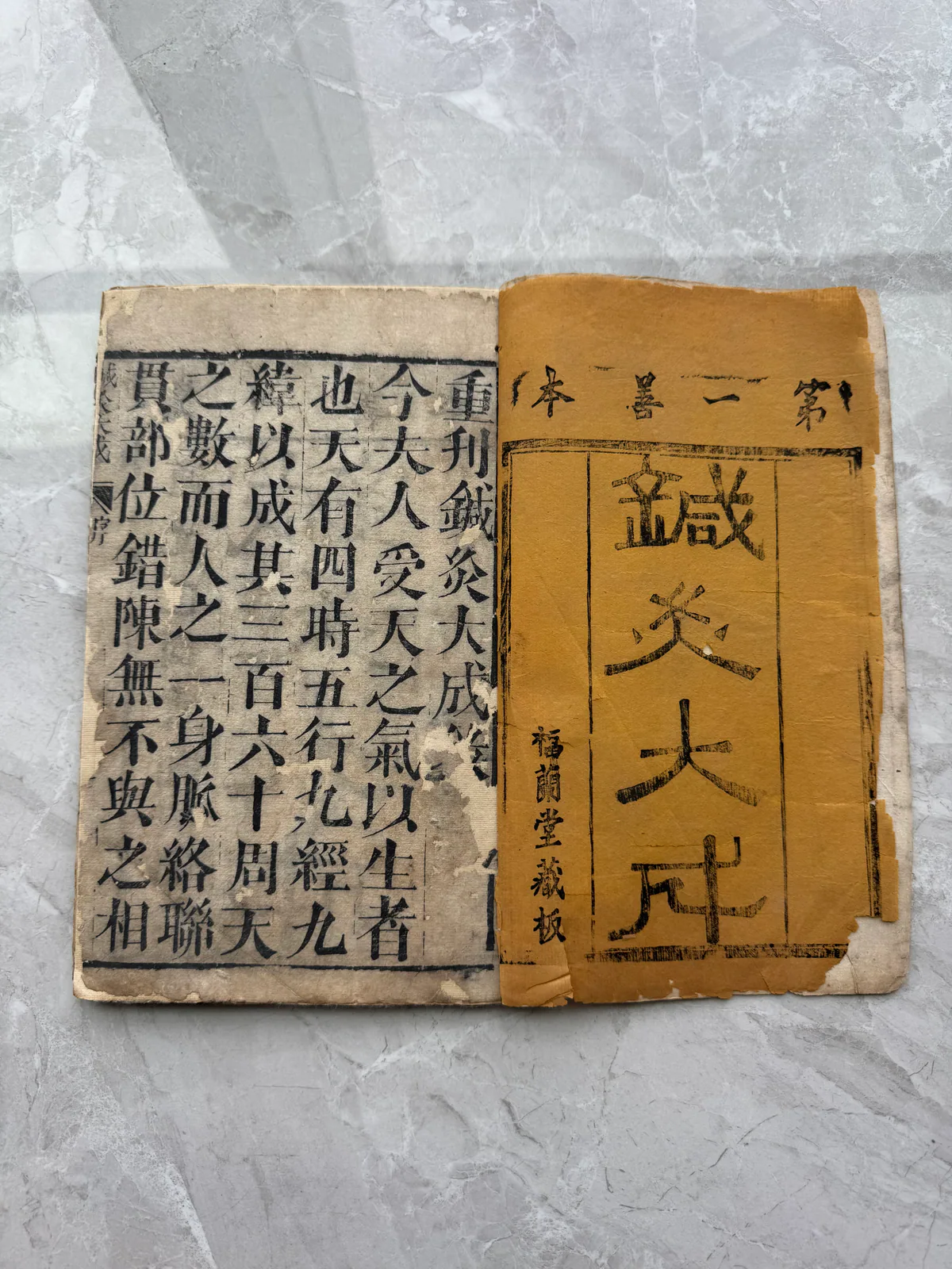这张图片呈现的是古籍《针灸大成》的封面及内页部分。封面为泛黄的旧纸张，上方是用篆体书写的书名“鍼灸大成”，侧面标注有“第二一卷”字样，另有“梅蘭堂藏板”的字样。下方露出的内页同样是泛黄的旧纸张，上面印着繁体汉字，内容可见“重刊鍼灸大成”“人之三百六十周”等与针灸相关的文字，整体纸张带有岁月留下的陈旧痕迹，对应文档中所描述的《针灸大成》古籍相关内容。