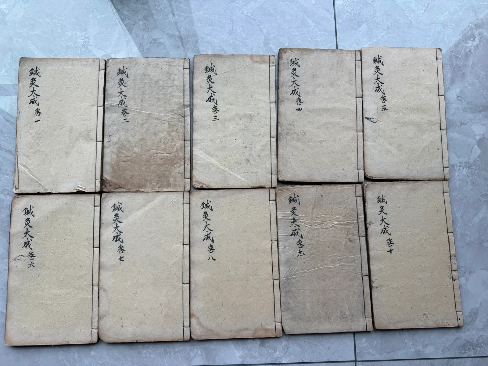 图片展示了郑福兰堂珍藏的《针灸大成》多卷本。共有九册，分别为卷一至卷九，每册封面均为浅棕色，书名及卷数以黑色字体印制。这些书册整齐排列，置于浅色地砖上。图片与上下文紧密相关，直观呈现了《针灸大成》这部典籍的分量，印证了其在中医史上的重要地位，以及郑福兰堂对这部典籍的珍视。