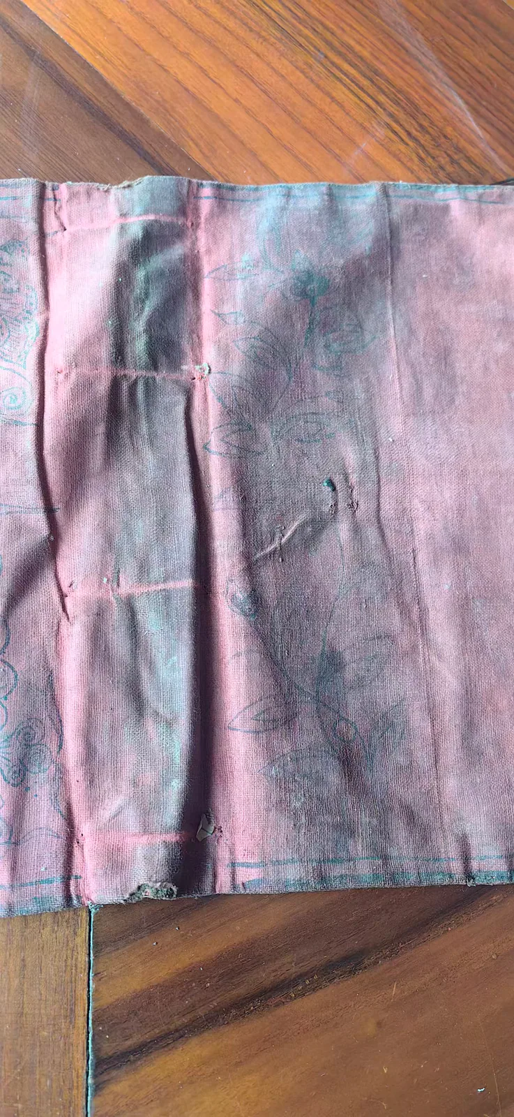 这张图片展示的是一个旧的针灸包特写，其包身主体为褪色的粉红色布料，布料表面因年代久远出现了明显的磨损痕迹，局部有发黑的污渍，还有细小的破损孔洞。包身布料上以深色线条绣绘有植物花卉纹样，左侧边缘还带有装饰性的图案，同时能看到用于固定针灸针的规整缝线痕迹。该针灸包是知识走向实践的见证者，包身的“郑福兰堂记”也藏着把针灸知识落到实际诊疗的承诺，那些原本仅在《针灸大成》上记载的穴位，都将通过这类针包转化为具体的实操内容。