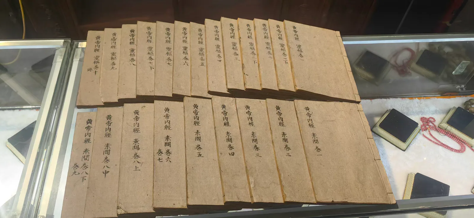 图片展示的是郑福兰堂珍藏版《黄帝内经》（线装多册），陈列于展柜中。书册整齐排列，封面为浅棕色，书名及卷数等信息清晰可见。部分书册上还贴有黄色标签，标注了书名和卷数。展柜内还摆放着其他古籍和文房四宝，如毛笔、砚台等，营造出浓厚的古籍文化氛围。此图与上下文紧密相关，直观呈现了《黄帝内经》的珍藏版样貌，呼应了文档中对《黄帝内经》作为中医理论基石的介绍。
