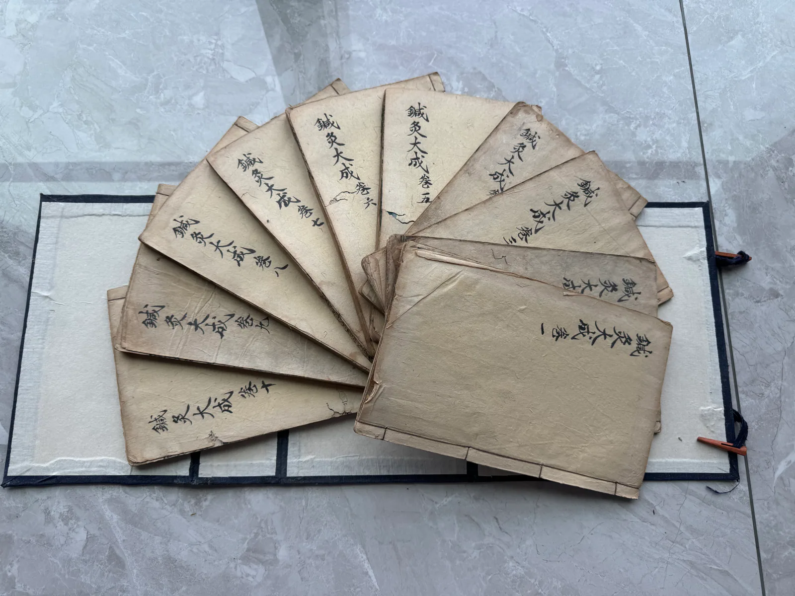 图片展示了郑福兰堂珍藏的《针灸大成》线装函套本。书本呈深蓝色布面，函套上贴有手书书签，古朴庄重。书本打开，内页书影清晰可见，每页书皮上均用黑色毛笔书写着“针灸大成”字样。这些书本整齐地堆叠在一起，部分书页微微卷曲，呈现出历史的痕迹。图片与上下文紧密相关，直观呈现了郑福兰堂对这套珍贵中医典籍的保存状态，体现了其对《针灸大成》的珍视。