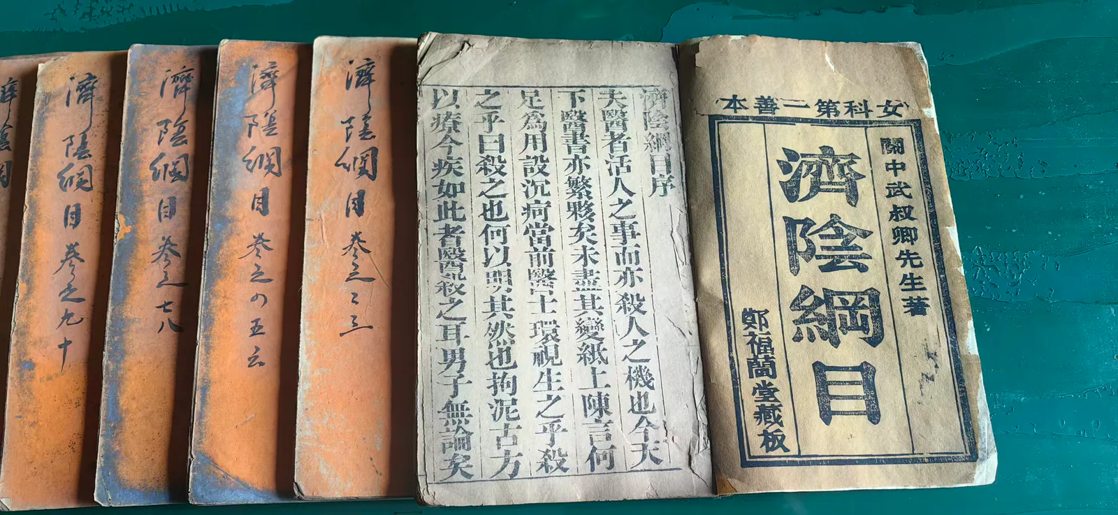 图片展示了郑福兰堂珍藏的《济阴纲目》多册线装书。书脊上用毛笔书写着书名及卷数，封皮为橙黄色，封面上有“郑福兰堂藏板”字样。其中一本封面上还印有“女科第一善本”“关中武叔卿先生著”等文字。这些书籍排列整齐，背景为绿色，整体呈现了郑福兰堂对这部女科典籍的珍藏状态，与上文提到的郑福兰堂关注妇科疾病、为患者提供专业诊疗依据相呼应。