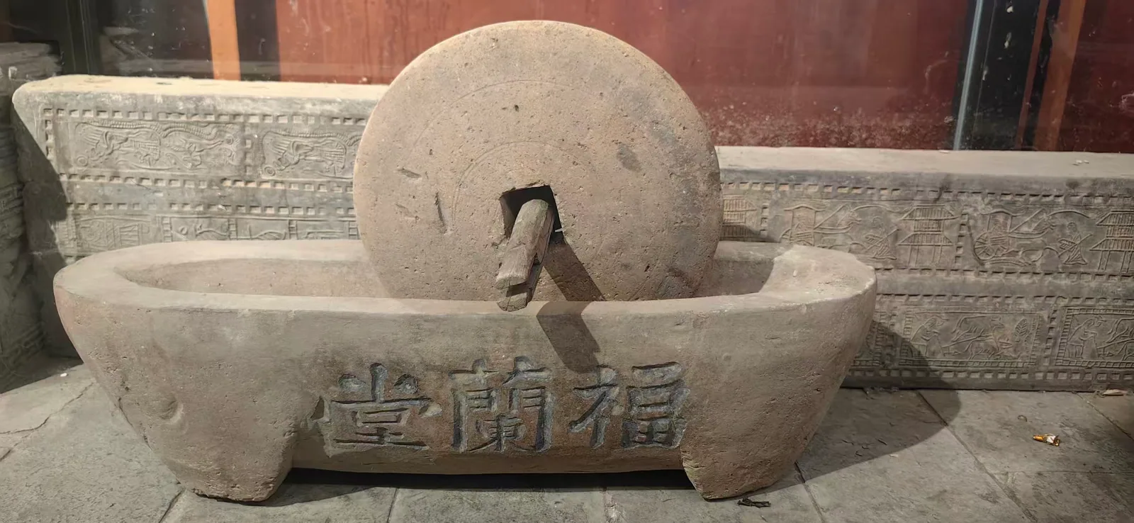 图片展示的是清朝刻有“福兰堂”药铺专用炮制的药碾。药碾由一个大石轮和一个石槽组成，石轮上刻有“福兰堂”字样，石槽底部也有相同字样。石轮与石槽之间有连接部分。背景是红木色的墙和一些石雕。该图片与上文提到的郑福兰堂将“大医精诚”过成生活日常的内容相关，直观呈现了福兰堂药铺专用炮制药材的工具，体现了其传统炮制工艺。