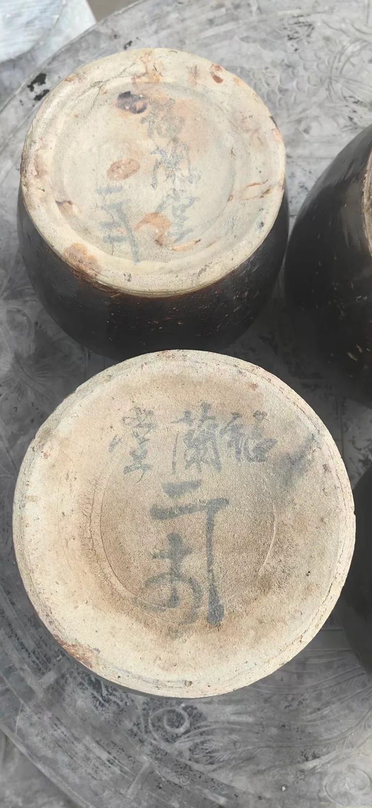 这张图片展示了两个瓷罐的底部，罐身为深色，底部胎体呈米白色，表面有磨损痕迹，其中一个底部有“郑福兰堂 三年”的字样，另一个底部写有“堂蘭福 二年”的字样。郑福兰堂是传统中医馆，这类器物是该医馆留存下来的药酒坛罐，印证了药酒需要用耐得住时间消磨的容器来盛装，符合药酒的制作特点，也对应了文档中关于郑福兰堂器物装着中医日常讲究的内容，这些坛罐见证了该医馆当年售卖、盛装药酒的日常场景。